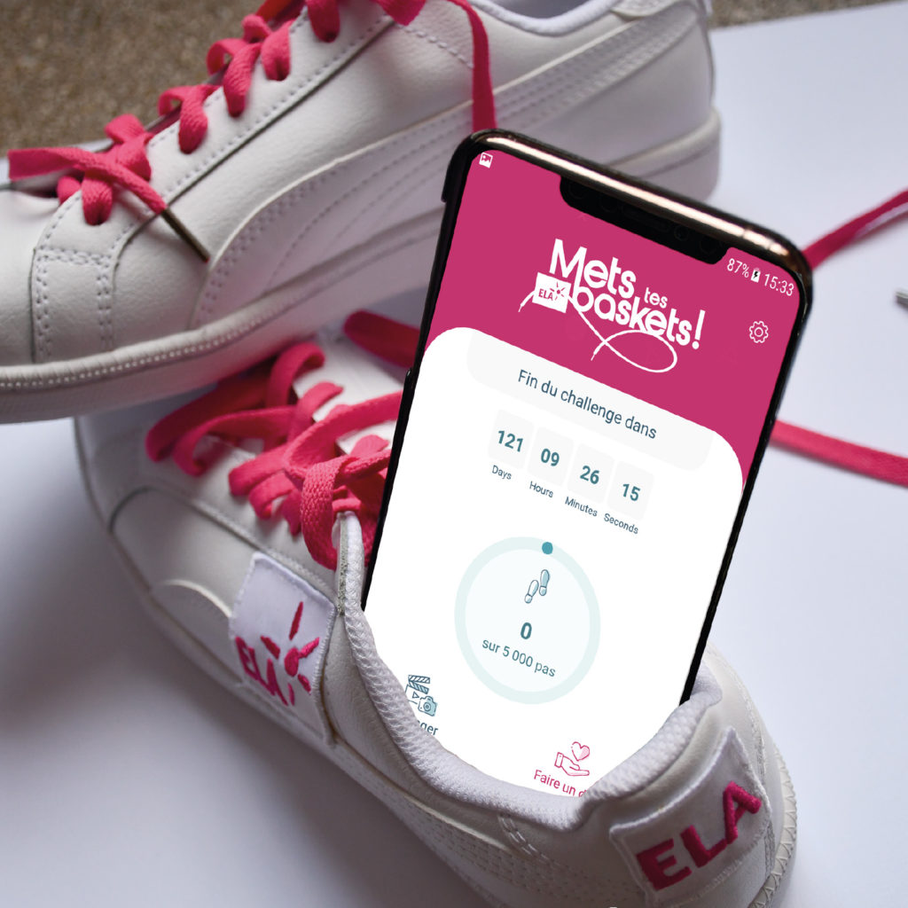 Smartphone affichant l'application Mets Tes Baskets, posé dans une basket blanche aux lacets roses marquée ELA