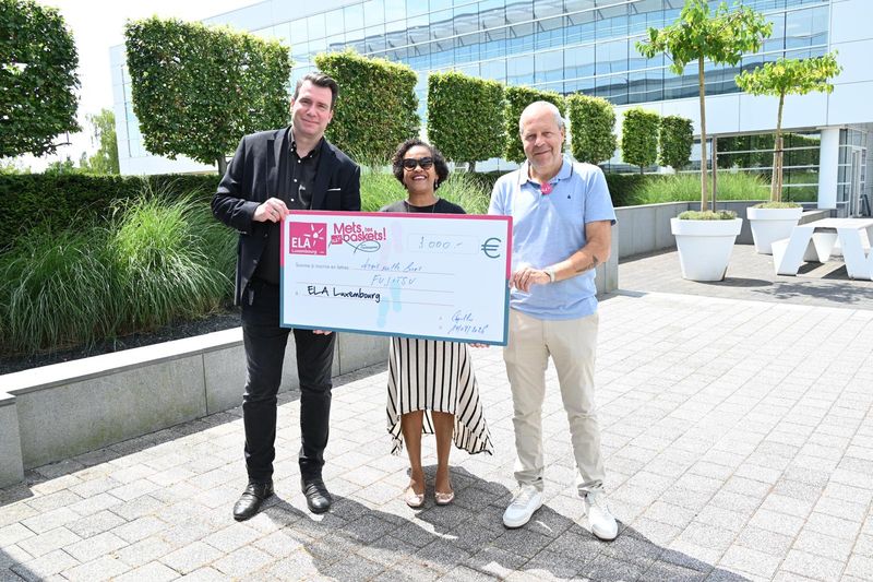 Trois personnes présentent le chèque « Mets Tes Baskets » de Fujitsu Luxembourg devant un immeuble de bureaux